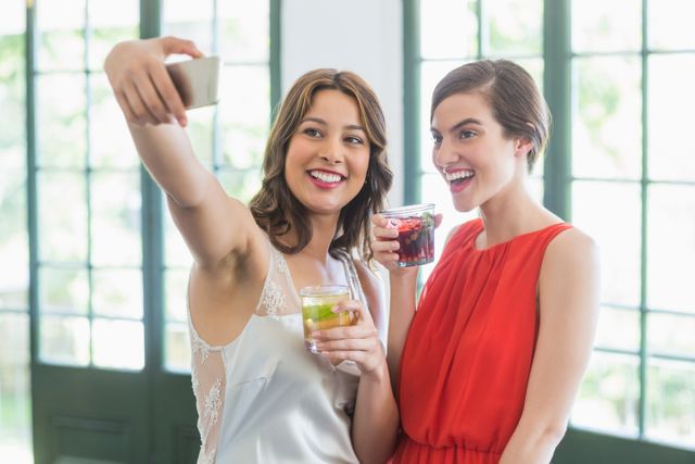 Two women are taking a selfie while holding cocktail glasses in a restaurant. They are smiling and appear to be enjoying their time together. This image can be used for promoting social events, restaurant advertisements, or lifestyle blogs focusing on friendship and leisure activities.