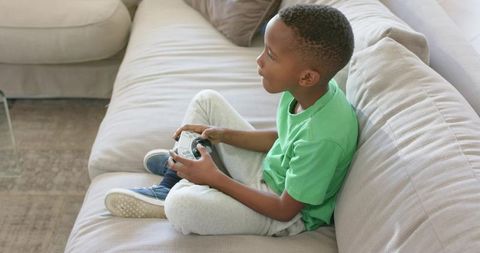 Black boy sitting on beige sofa playing video game with wireless controller at home