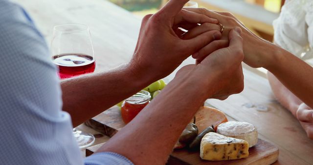 A Caucasian couple engages in a romantic gesture, holding hands over a table adorned with wine and cheese, with copy space. Their intimate moment is accentuated by the warm setting, suggesting a celebration or a special date.