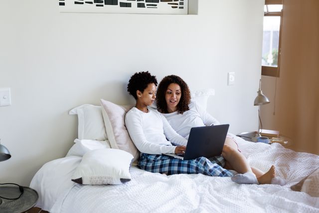 Happy, diverse lesbian couple sitting on bed looking at laptop together. Togetherness, free time and domestic life concept.
