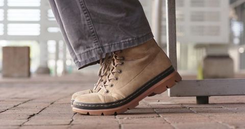 Asian man balancing on toes in tan lace-up boots on brick pavement near bench