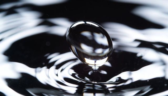 Hovering transparent water droplet above dark pool with concentric ripples and reflection