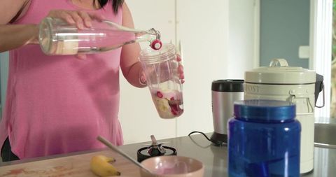 Mature woman pouring water into blender cup with banana and yogurt making healthy smoothie