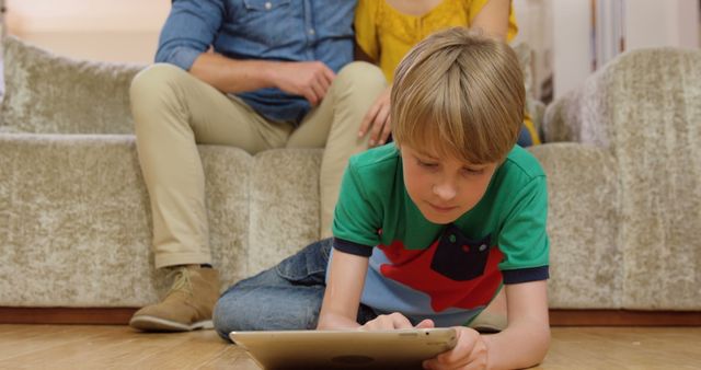 Young boy lying on floor using a tablet, with parents sitting in background. Depicts modern family lifestyle and child's engagement with technology. Useful for themes on digital learning, parenting, modern technology, and family activities.