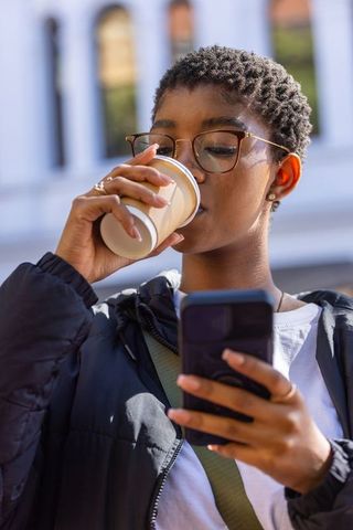 African american woman drinking coffee checking phone on city street wearing eyeglasses