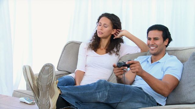 Couple lounging on sofa with man playing video games and woman appearing bored, suggesting relaxed but unequal participation in entertainment. Useful for illustrating relationship dynamics, leisure activities, or video game culture from opposing perspectives.