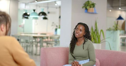African american woman sitting on pink sofa talking with colleague in coworking lounge