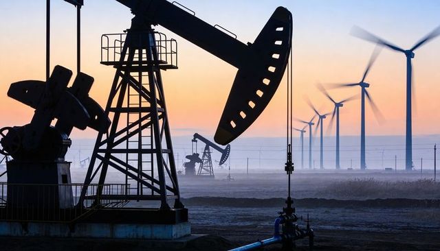 Pumpjack silhouetted while wind turbines turning at sunrise over misty oilfield horizon