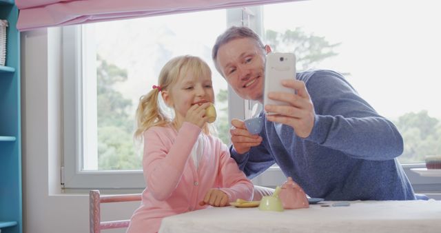 Father and daughter wearing casual clothing enjoying a playful tea party at home and taking a selfie together. Could be used to illustrate family bonding, happy childhood, or modern family life concepts. Perfect for advertisements related to parenting, family activities, or lifestyle blogs.