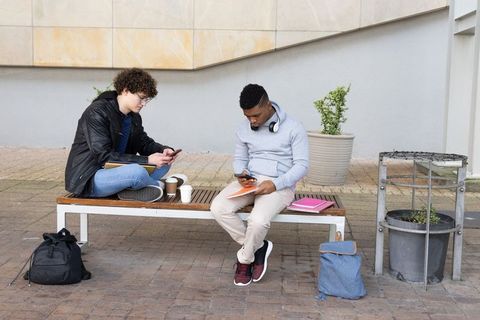 College students studying outdoors on campus bench using smartphones and notebooks