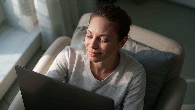 Calm woman working on laptop in armchair near sunlit window, minimalist home workspace