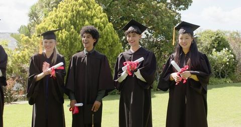 Diverse graduates celebrating outdoors in mortarboards holding diplomas with red ribbons