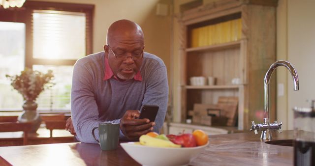 Senior man seen using a smartphone while leaning in kitchen. He is surrounded by a fruit bowl and coffee mug, suggesting a relaxed, homey atmosphere. Useful for themes on technology adoption among elderly, home lifestyle, casual living, and everyday activities.