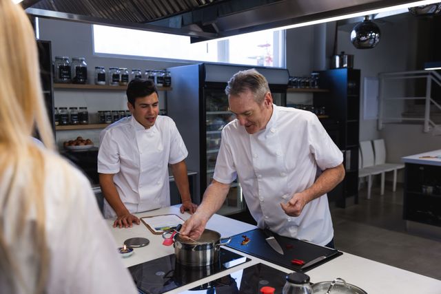 Multi ethnic group of chefs cooking in a modern busy kitchen, male chef checking a pan and instructing students. Cookery class at a restaurant kitchen. Workshop cooking food.
