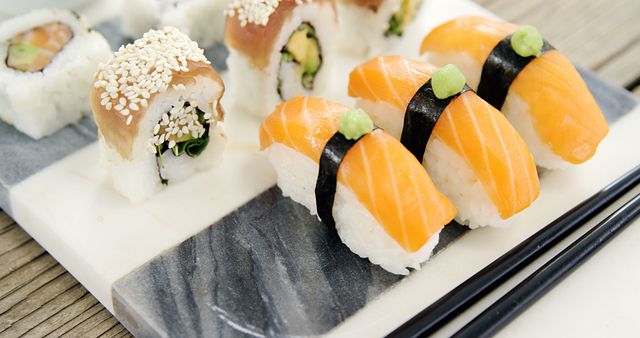 A variety of sushi, including nigiri topped with salmon and maki rolls sprinkled with sesame seeds, is presented on a marble plate, with chopsticks alongside. Sushi is a traditional Japanese dish enjoyed worldwide for its fresh flavors and artistic presentation.