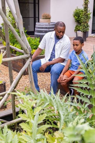 African american father comforting preteen son while sitting on planter in courtyard garden