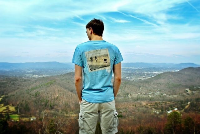 Man standing on a hilltop, enjoying an expansive view of mountains. He is wearing a blue t-shirt and khaki shorts, gazing serenely into the distance. Ideal for concepts of relaxation, outdoor activities, serene getaways, and contemplating nature's beauty.