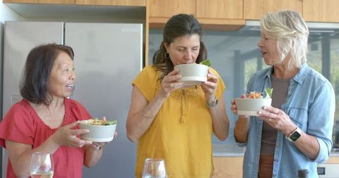 Three friends gathering in kitchen sharing fresh salad bowls and lively conversation