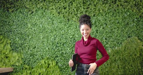 African American woman smiling and holding tablet by living wall in burgundy sweater
