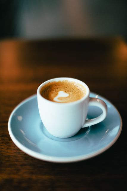 This image shows a cup of espresso placed on a blue saucer with foam art in the shape of a heart, set on a wooden table. Suitable for illustrating coffee shop menus, blog posts about coffee, or advertisements for cafés.
