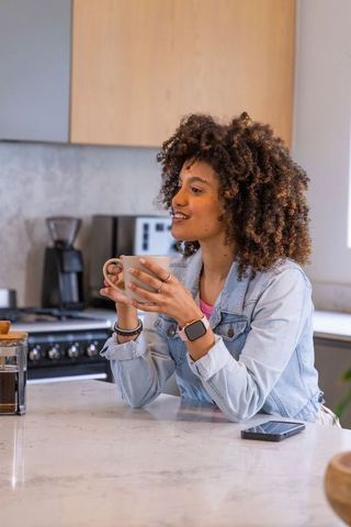 African american woman sipping coffee at kitchen counter in denim jacket on sunlit morning
