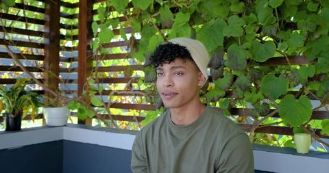 Young man wearing beanie sitting on sunlit patio bench among lush green vines holding cup