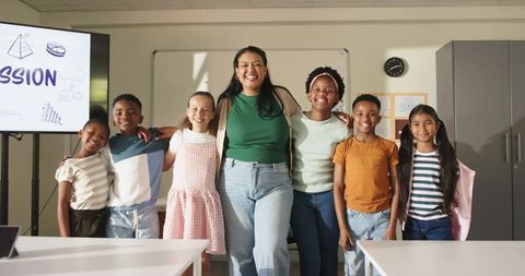 Smiling teacher and diverse elementary students standing arm in arm in bright classroom