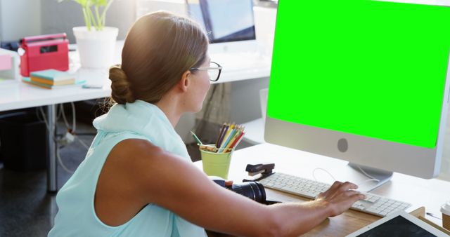 An image showing a graphic designer sitting at her desk in a well-lit office environment, working on a computer with a green screen. This can be used for illustrating concepts related to creativity, graphic design, professional environments, office settings, and workplace technology.