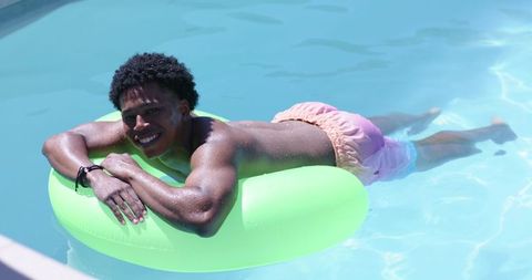 Smiling young man floating on neon tube in sunlit backyard pool for summer leisure