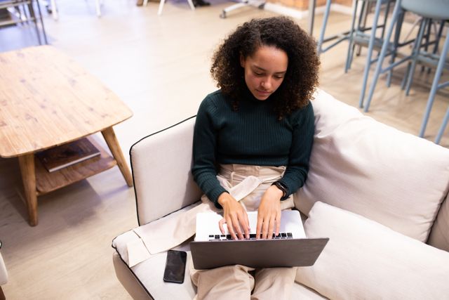 Biracial businesswoman sitting on a sofa in a modern creative office, working on a laptop. Ideal for use in articles about remote work, modern office environments, productivity, and professional women in tech. Suitable for business and technology blogs, workplace culture features, and promotional materials for coworking spaces.