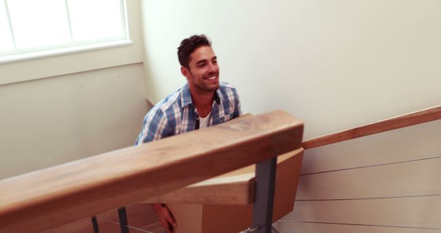 Man carrying cardboard box up stairs, dressed casually in plaid shirt and smiling. This photo can be used for topics related to moving into a new home, relocation, residential life, logistics, effortful tasks, and youthful energy. Ideal for real estate brochures, moving service advertisements, lifestyle blogs, and articles about the moving process.