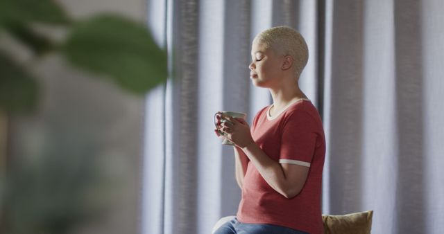 Female with short blond hair holding coffee cup in hands, sitting comfortably by window. Could be used for well-being content, meditation or relaxation promotions, lifestyle blogs depicting moments of peace and contemplation, or advertisements focused on home comforts or morning routines.
