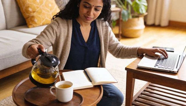 Woman pouring tea while checking laptop and writing notebook in cozy home workspace
