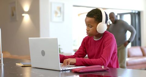 African american father supervising son typing on laptop at sunlit kitchen island