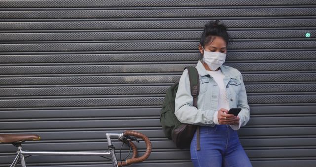 Young woman wearing mask, leaning against metal shutters, using smartphone. Bicycle next to her. Suitable for themes related to urban living, safety measures, modern lifestyle, and transportation.