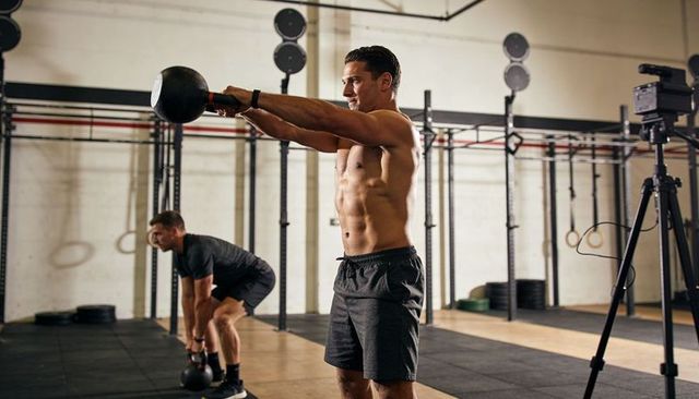 Shirtless athlete performing kettlebell swing in industrial crossfit gym trainer filming