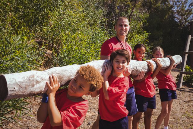 Trainer helping a group of children carry a heavy wooden log during an outdoor obstacle course. Ideal for use in articles or advertisements about fitness, teamwork, children's activities, summer camps, and outdoor education programs.