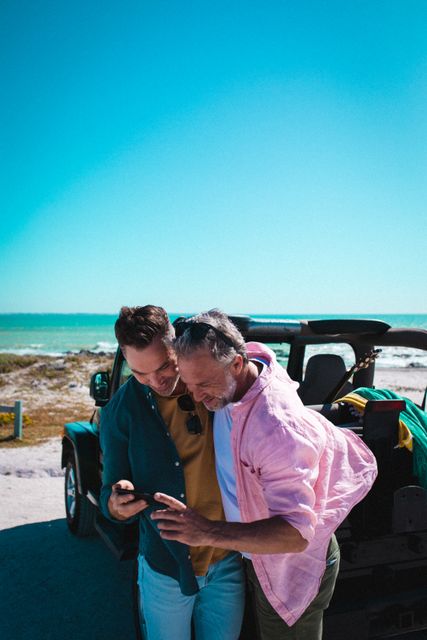 Two friends standing by an off-road vehicle at the beach, sharing a moment while looking at a smartphone. Ideal for themes related to summer vacations, road trips, friendship, outdoor adventures, and technology use during travel.