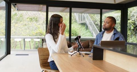 Diverse co-hosts recording conversational podcast at bright glass-walled home studio