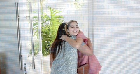 Two women hugging at home entrance welcoming reunion with bags and sunlit glass door