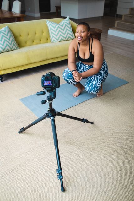 African American female blogger recording a fitness video on a digital camera while sitting on a yoga mat in a modern living room. Ideal for content related to fitness, healthy lifestyle, home workouts, social media influencers, and vlogging.
