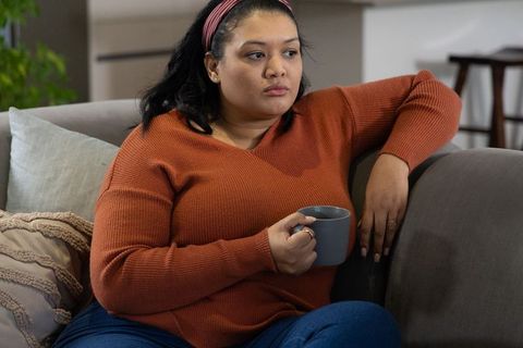 Pensive woman relaxing on sofa with mug wearing rust sweater and blue jeans at home
