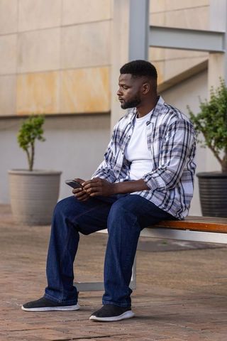 African american man sitting on bench holding smartphone looking thoughtful in urban plaza