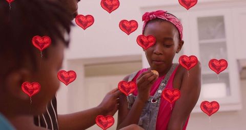 Young girl tasting cookie in kitchen with supportive hand and red heart balloons