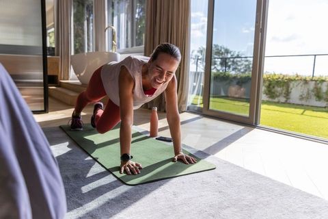 Woman holding plank and smiling on yoga mat in sunlit bedroom with garden view