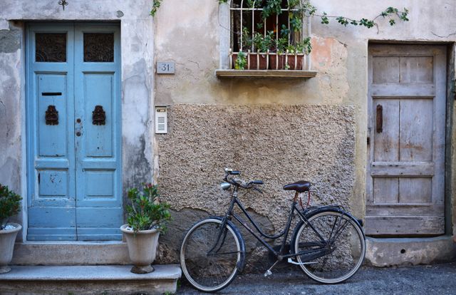 Vintage bicycle resting against rustic Italian building with blue door, potted plants, and weathered textures. Ideal for storytelling, travel websites, cultural blogs, and historical preservation articles.