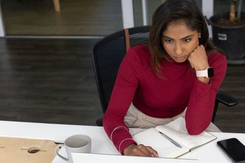 Indian professional woman working on laptop at modern minimalist office desk, concentrating on notes