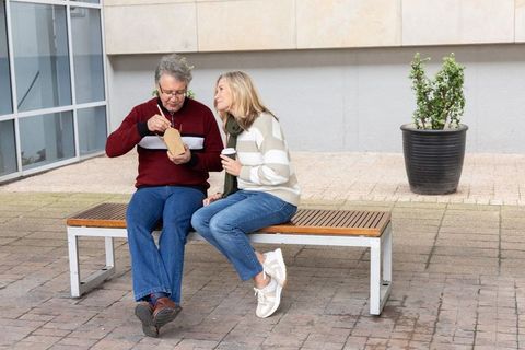 Senior couple sitting on bench sharing takeaway and coffee in modern urban plaza