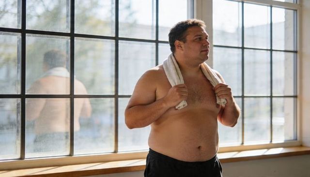 Standing shirtless man holding towel by grid window reflecting post-workout calm in morning sunlight