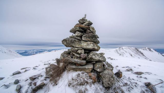 Stone cairn on snowy mountain ridge with lichen-covered rocks and distant blue peaks
